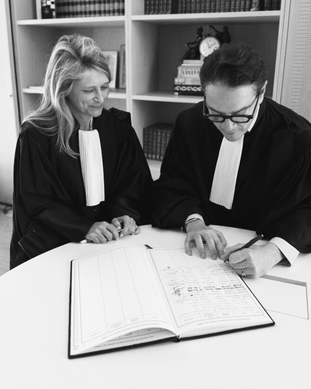 David Koubbi et Virginie Ricaud, en robe d’avocat, penchés sur un registre ouvert, en noir et blanc